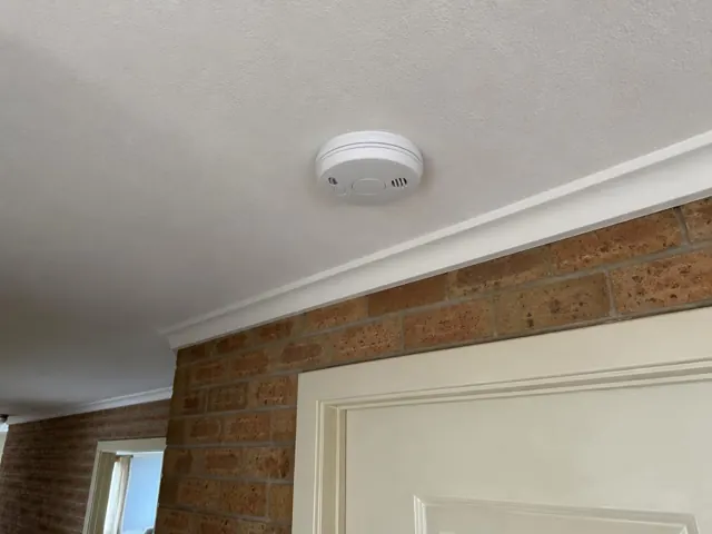 Ceiling-mounted smoke alarm above a doorway in a brick home interior in Woongarrah